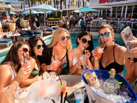 Five young women in sunglasses enjoy cocktails by a pool at a sunny outdoor venue, with a "Rose" phone case visible. They are laughing and taking a selfie, with bottles of Tito's and Patron in a cooler in the foreground.