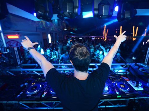 DJ with arms raised in celebration at a vibrant nightclub, surrounded by bright blue stage lights and a cheering crowd.