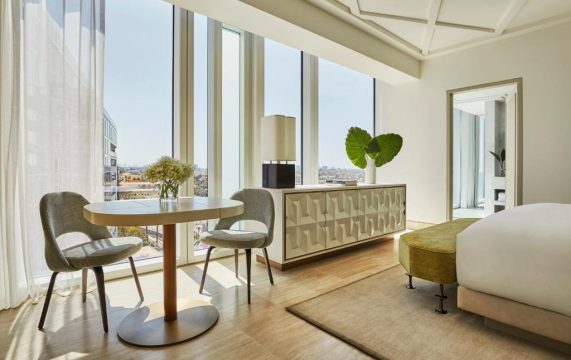 Modern hotel room with a round dining table and two chairs near a large window, featuring a contemporary dresser and a plush bed with a green ottoman.