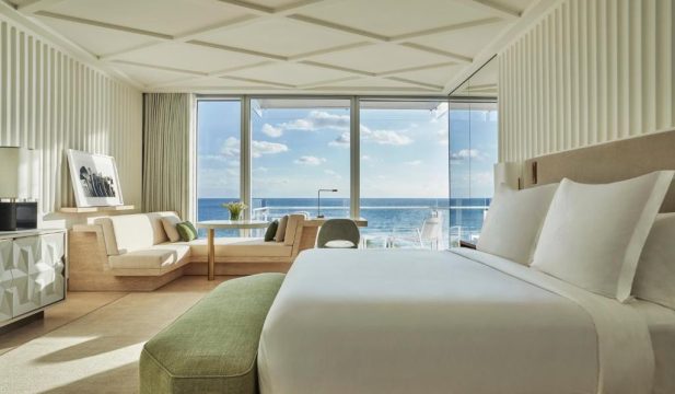 A luxurious hotel suite with a king-size bed, a modern sofa, and a desk, featuring a stunning ocean view through floor-to-ceiling windows. The room is decorated in neutral tones with accents of green, creating a serene and elegant ambiance.