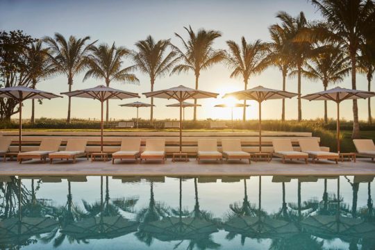 Luxury resort poolside with lounge chairs and umbrellas at sunset, palm trees and ocean in the background. Perfect for a tropical vacation.