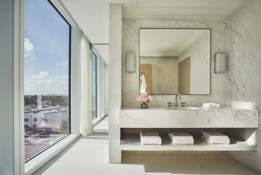 Luxurious marble bathroom with a large mirror, vanity, and fluffy white towels, featuring a stunning panoramic city view through floor-to-ceiling windows.