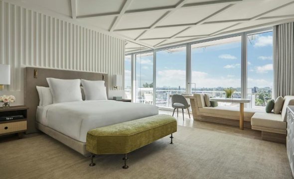 Luxurious hotel suite with a king-size bed, plush white bedding, and a modern design featuring floor-to-ceiling windows offering panoramic views of a sunny, tree-lined cityscape.