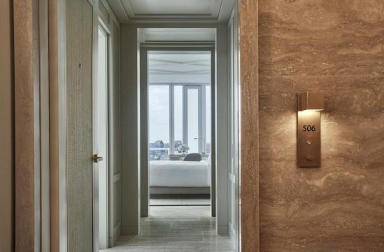 Luxury hotel hallway with a modern door number sign "506" on textured marble wall, leading to a bright bedroom with a large window and city view.
