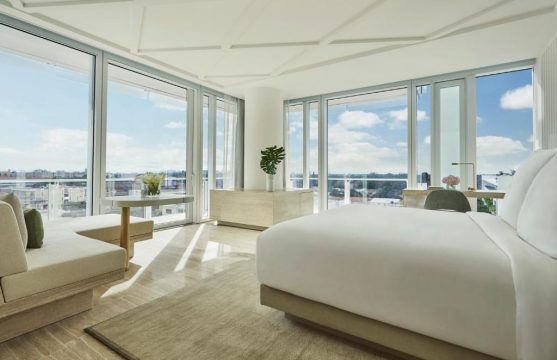 Luxurious hotel suite with floor-to-ceiling windows offering panoramic city views, a king-sized bed, modern seating area, and a minimalist decor.