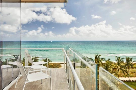 A modern balcony with white chairs overlooks a turquoise ocean and sandy beach lined with palm trees under a cloudy blue sky.
