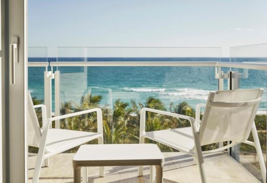 Two white chairs and a small table on a balcony overlooking a turquoise ocean and palm trees, offering a serene beach vacation view.