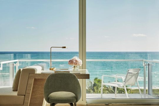 A modern hotel room desk with a lamp and flowers overlooks a bright blue ocean and balcony with white chairs, offering a serene coastal view.
