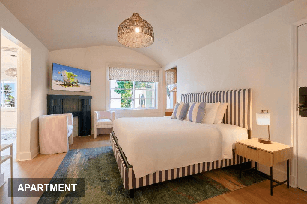 A spacious apartment bedroom featuring a king-size bed with a striped headboard, a large window with sheer curtains, and a TV mounted above a fireplace.