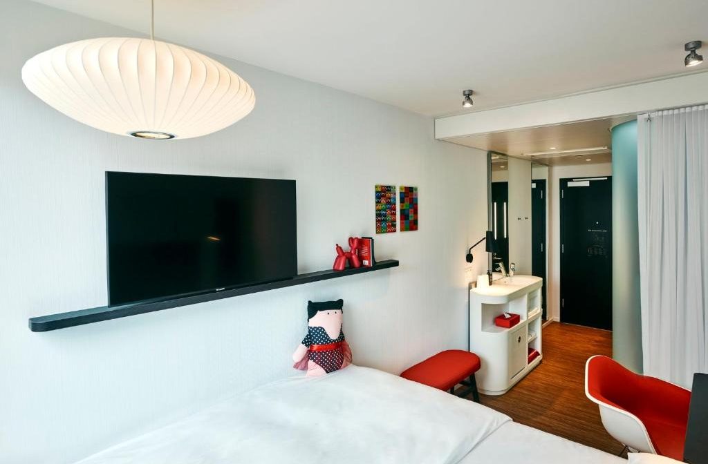 Modern hotel room with a large flat-screen TV mounted on a black shelf above a bed, a white pendant light, and a small red stool.