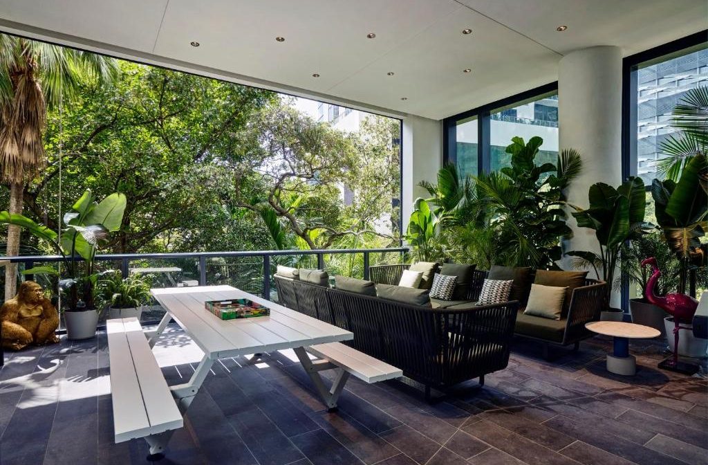 Modern outdoor patio with a white picnic table and a dark wicker sofa set, surrounded by lush tropical plants and city buildings visible through the greenery.