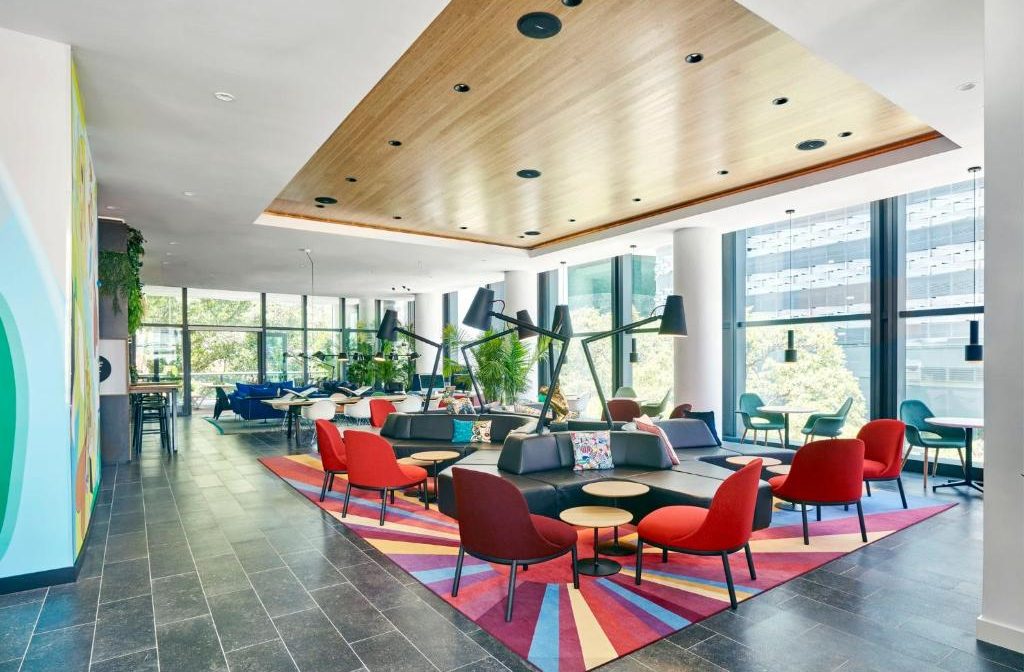 Modern, vibrant coworking space with colorful geometric rug, red chairs, and large windows overlooking a city.