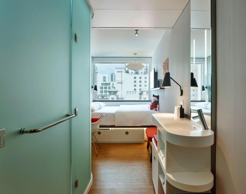 Modern hotel room with a compact bathroom featuring a white sink and vanity, leading to a bedroom with a twin bed, desk, and large window overlooking a city skyline.