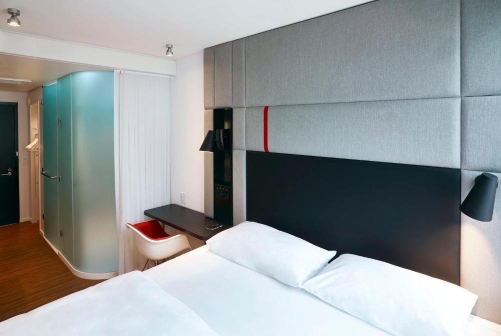 Modern hotel room with a king-size bed, a sleek black headboard, and a minimalist desk with a red chair. A frosted glass shower enclosure is visible to the left.