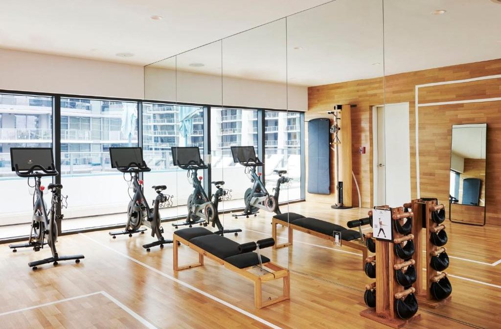 Modern home gym with multiple spin bikes, weight benches, and kettlebells, featuring large windows with city views and mirrored walls reflecting the polished wood interior.