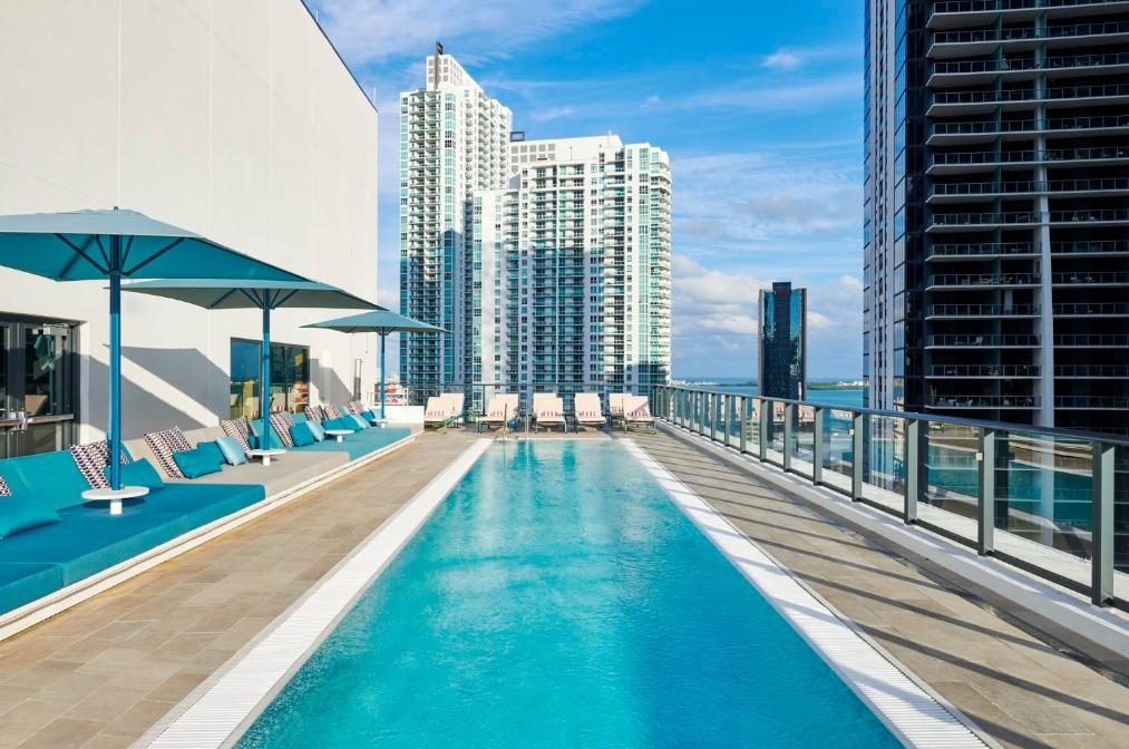 Rooftop infinity pool with turquoise water, surrounded by modern lounge seating and umbrellas, with a backdrop of towering city skyscrapers under a bright blue sky.