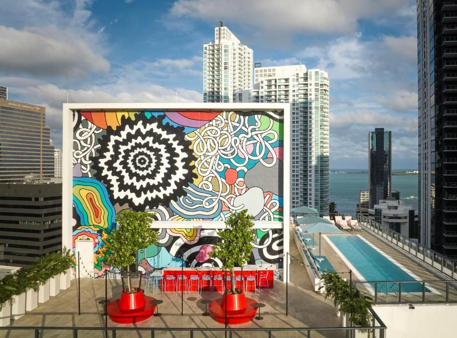 Rooftop bar in Miami with a vibrant mural, pool, and city skyline. Modern outdoor seating and lush greenery complement the urban oasis.