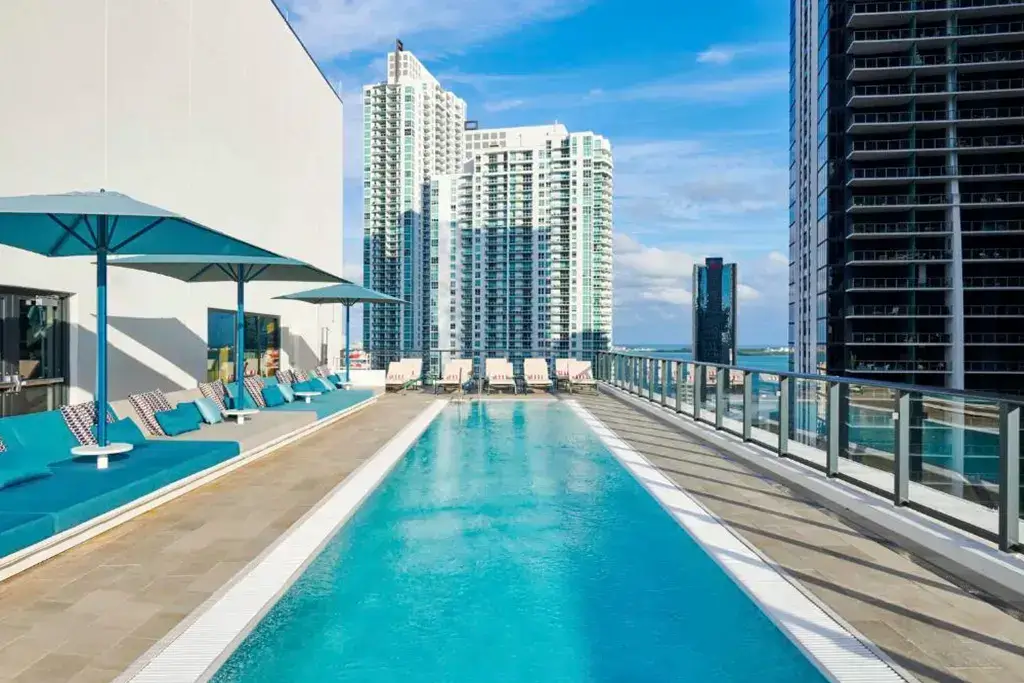 A vibrant rooftop pool with turquoise lounge chairs and umbrellas offers stunning city views, with skyscrapers and a bright blue sky in the background.