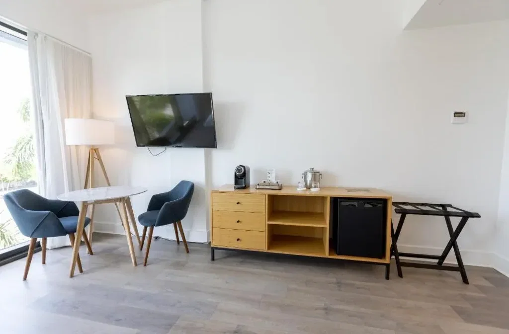 A modern hotel room sitting area features a flat-screen TV, a small round dining table with two blue chairs, and a wooden buffet with a mini-fridge and coffee maker.