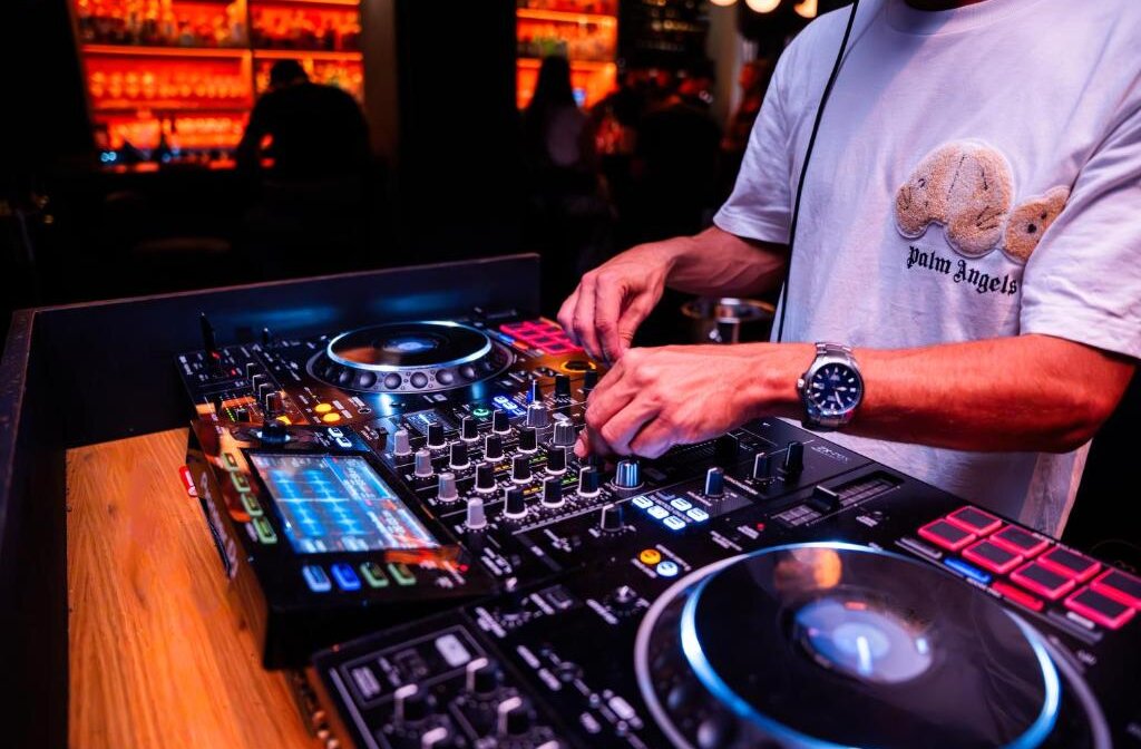 DJ's hands mixing music on a professional DJ controller with illuminated decks and a screen displaying waveforms, in a dimly lit club with a bar in the background.