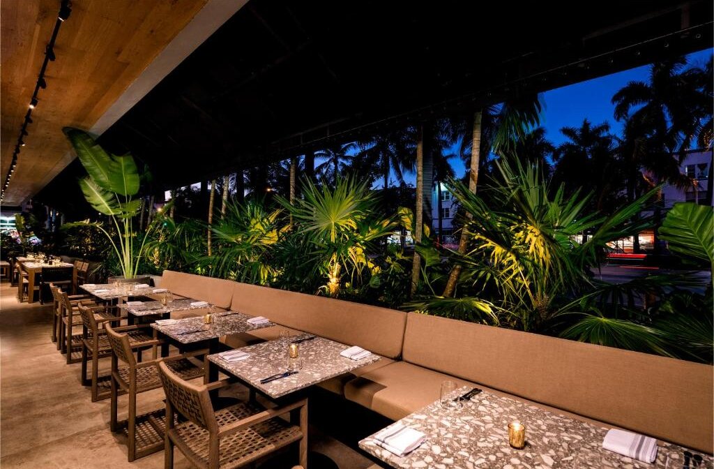 Outdoor dining at a tropical restaurant at dusk, with palm trees, lush greenery, and tables set for dinner.