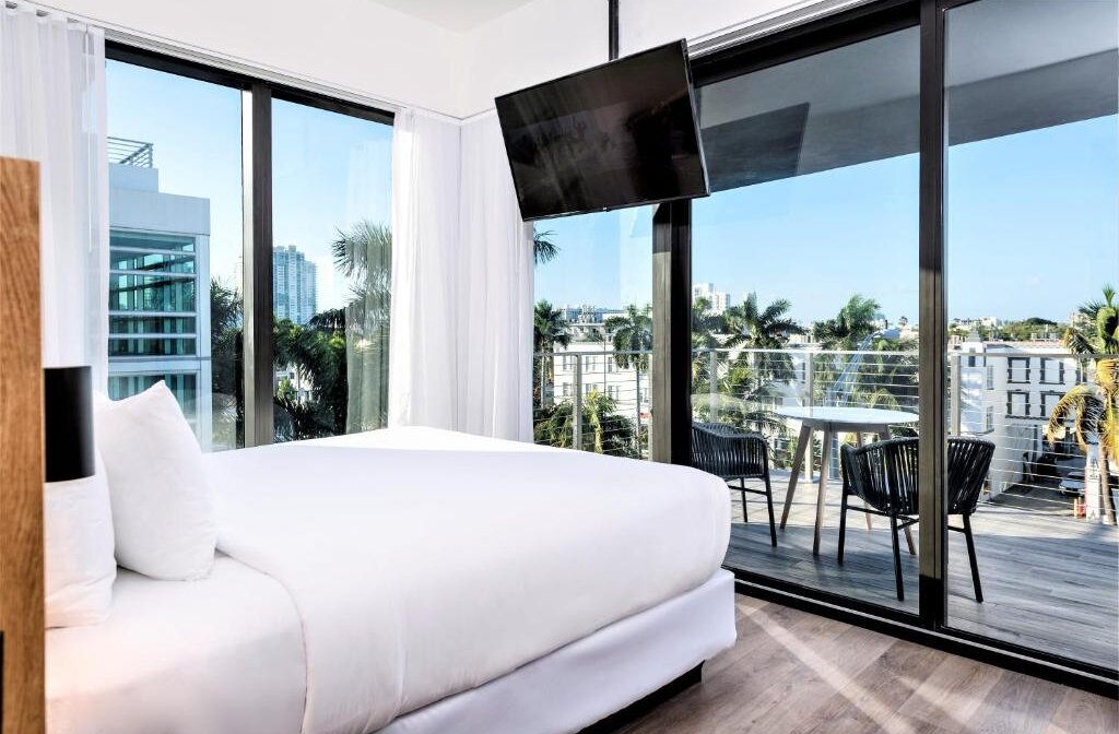 Modern hotel bedroom with a plush white bed, large windows opening to a balcony with city and palm tree views, and a wall-mounted TV.