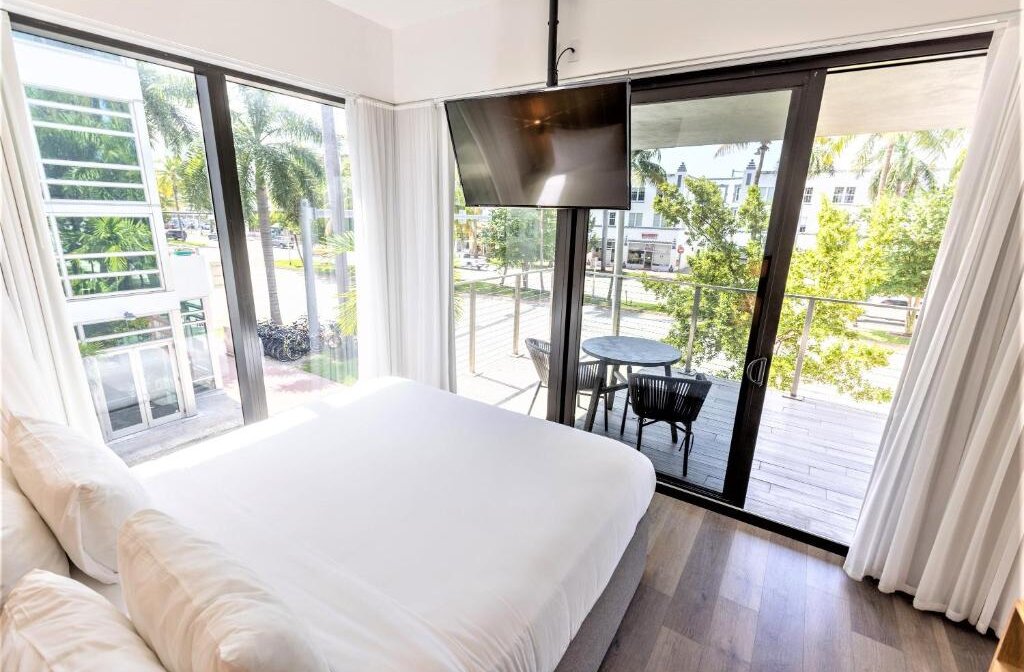 Modern hotel room with a king-size bed, large windows, and a private balcony overlooking palm trees and a street scene.