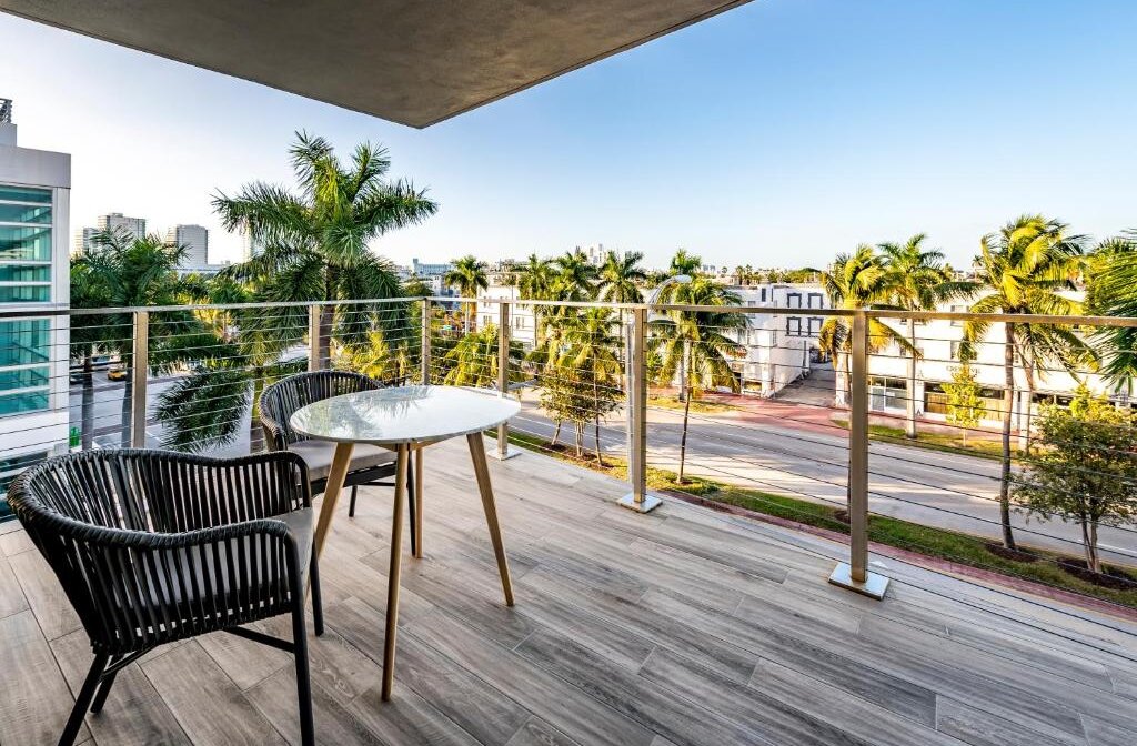 A modern balcony with a small table and chairs overlooks a street lined with palm trees and apartment buildings, bathed in warm sunlight.